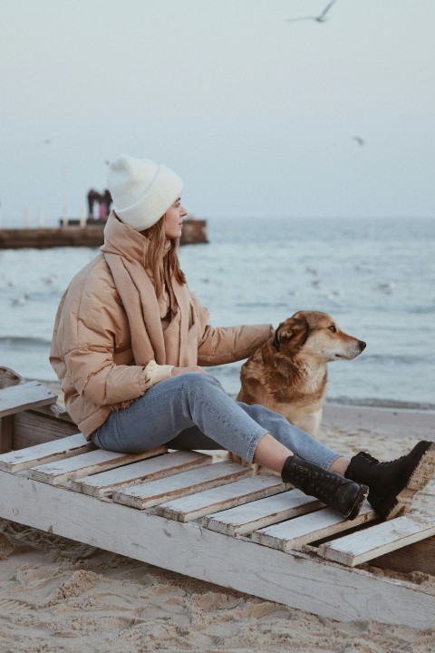 woman in brown jacket and blue denim jeans sitting on brown wooden bench during daytime Dz